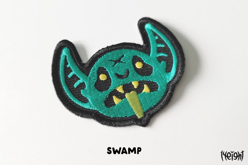 Pu&ograve; includere: Toppa ricamata verde e gialla di un mostro dei cartoni animati con la scritta "SWAMP" sotto.