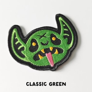 Pu&ograve; includere: Toppa ricamata verde di un mostro dei cartoni animati con un ghigno pieno di denti e una lingua rosa che sporge. Il mostro ha grandi orecchie e occhi gialli.