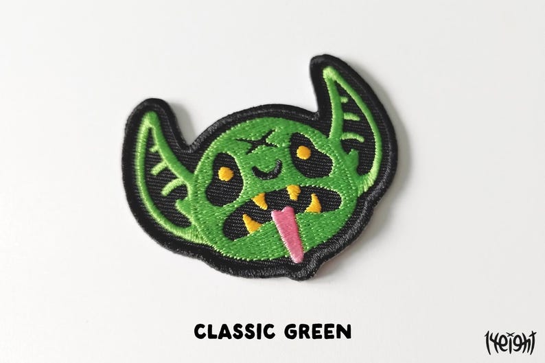 Pu&ograve; includere: Toppa ricamata verde di un mostro dei cartoni animati con un ghigno pieno di denti e una lingua rosa che sporge. Il mostro ha grandi orecchie e occhi gialli.