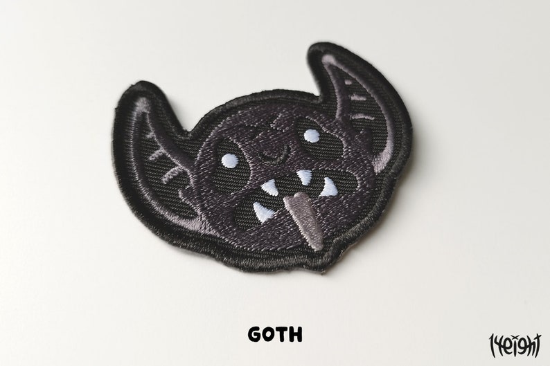 Pu&ograve; includere: Toppa da stirare ricamata nera di un pipistrello dei cartoni animati con occhi e denti bianchi. La toppa dice "GOTH" in basso.