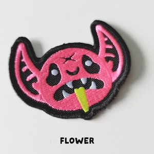 Pu&ograve; includere: Toppa ricamata rosa con un mostro dei cartoni animati con occhi neri, una lingua verde che sporge e la parola "FLOWER" sotto.