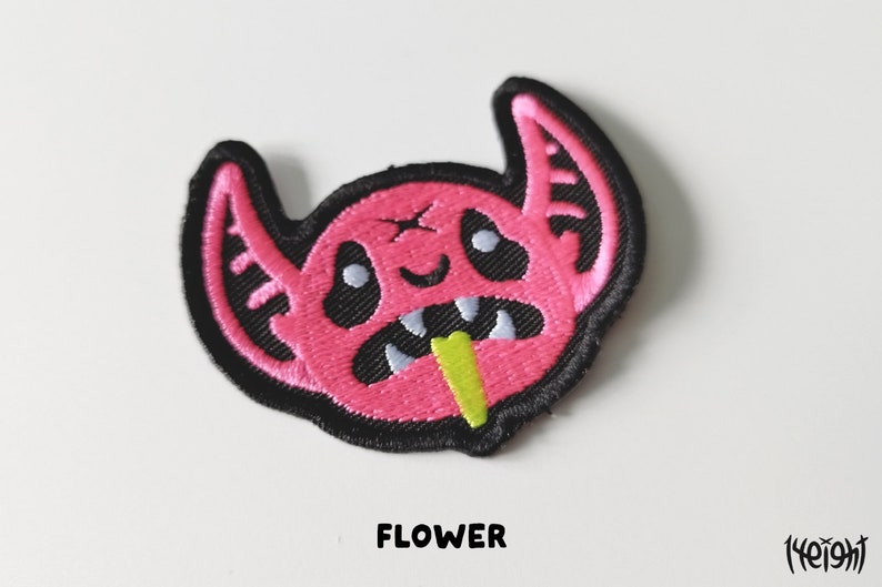 Pu&ograve; includere: Toppa ricamata rosa con un mostro dei cartoni animati con occhi neri, una lingua verde che sporge e la parola "FLOWER" sotto.