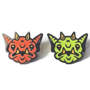 May include: Two enamel pins shaped like stylized creature heads with pointed ears and horns. One pin is orange with black outlines and yellow accents, while the other is lime green with black outlines and yellow accents. The text 14eight is visible.