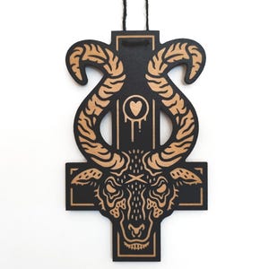 Op de afbeelding: Zwarte en bruine houten wanddecoratie met een gestileerde geitenkop in een kruisvorm. Het ontwerp bevat een hart en druipende details. Het stuk wordt opgehangen met een zwart touwtje.