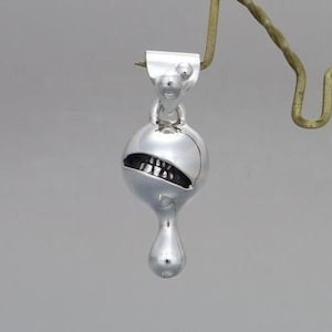 May include: A silver-colored pendant with a unique design, featuring a spherical body with a wide, toothy grin and a teardrop-shaped bottom. The pendant is suspended from a small, stylized head and is displayed on a decorative metal stand.