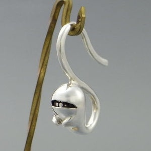 Könnte beinhalten: Ein einzelner silberfarbener Ohrring mit einem einzigartigen Design. Der Ohrring hat eine abgerundete, bauchige Form mit kleinen, dunklen Akzenten, die möglicherweise Augen oder einen Mund darstellen. Der Ohrring hängt an einem gebogenen Haken.