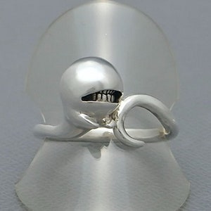 Könnte beinhalten: Ein silberner Ring mit einem stilisierten Kreaturendesign. Der Ring hat einen abgerundeten Kopf mit einem zahnigen Grinsen und ein gebogenes Band. Der Ring wird auf einem transparenten, kegelförmigen Ständer präsentiert.
