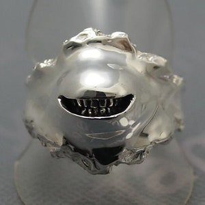 May include: A silver ring with a unique design. The ring features a textured, organic shape with a smiling face carved into the front. The mouth is open, revealing a row of teeth. The ring is displayed on a clear stand.