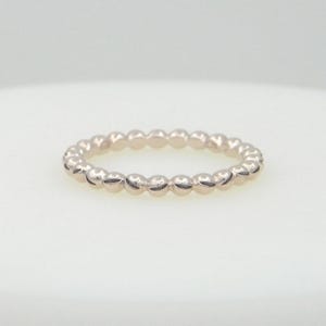 Peut inclure: Une bague en argent avec un motif de perles. La bague est circulaire et comporte de petites perles espacées uniformément sur tout le tour. La bague est posée sur un fond blanc.