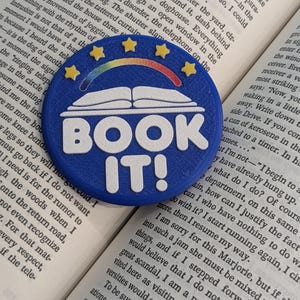 May include: A circular blue badge featuring the white text BOOK IT! below a white open book icon. A rainbow arch sits above the book, flanked by four small yellow stars. The badge is textured and rests on an open book with visible printed text.