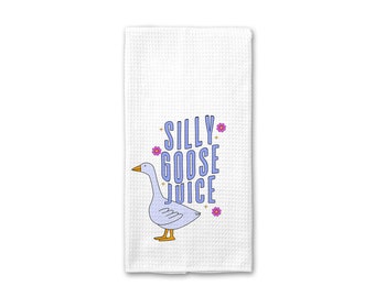 Könnte beinhalten: Ein weißes Küchentuch mit einer blauen Gänseillustration und dem Text "Silly Goose Juice" in lila Buchstaben. Rosafarbene Blumen sind um die Gans und den Text verstreut.