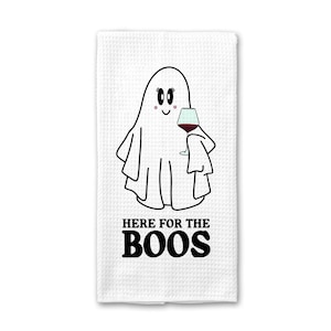 May include: White waffle-weave kitchen towel with a cartoon ghost holding a wine glass. The ghost has a friendly expression and is outlined in black. The text "HERE FOR THE BOOS" is printed in black block letters below the ghost.