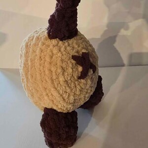 May include: A round, cream-coloured crocheted plush toy featuring dark brown accents on the top, bottom, and sides. The plush has a textured, soft yarn surface with a dark brown cross-shaped detail stitched onto the side of its spherical body.