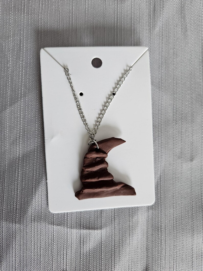 May include: A brown ceramic Sorting Hat pendant on a silver chain necklace. The pendant is shaped like the iconic hat from the Harry Potter series.