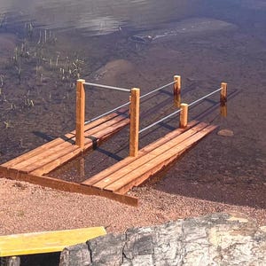 May include: A wooden dock extends into the water, featuring a rectangular platform with wooden planks. Vertical wooden posts support horizontal metal bars. The dock is partially submerged in the water, with a sandy shoreline and a yellow bench in the foreground.