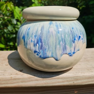 May include: A ceramic jar with a lid, featuring a rounded shape and a glossy finish. The jar is primarily off-white with a blue and green watercolor-like design around the middle. The lid is a matching off-white color.