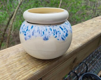 May include: A ceramic jar with a lid. The jar is off-white with blue speckled accents around the middle. The lid is a matching off-white color. The jar is sitting on a wooden surface.