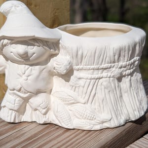 May include: A white ceramic planter featuring a smiling scarecrow figure leaning against a wheat sheaf design. The planter has a round opening and is decorated with corn and rope details, perfect for fall decor.