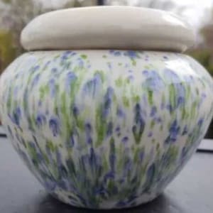 May include: A ceramic jar with a lid. The jar is white with a pattern of blue and green brushstrokes. The lid is a solid, off-white color. The jar has a rounded shape.