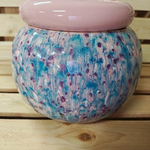 May include: A round ceramic jar with a lid. The jar has a speckled pattern of blue, purple, and white on a light pink base. The lid is a solid light pink color. The jar is sitting on a wooden surface.