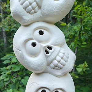 May include: A stacked sculpture of three white skulls, each with large eye sockets and a grinning mouth. The skulls are arranged vertically, creating a whimsical and slightly spooky decorative piece. The sculpture is set against a backdrop of green foliage.