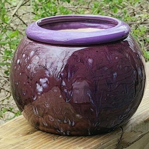 May include: A round, glazed ceramic pot with a deep purple hue and a glossy finish. The pot features a lighter purple rim and a textured surface with white speckles. The pot is sitting on a wooden surface.