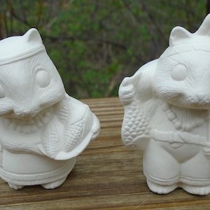 May include: Two unpainted ceramic figurines of anthropomorphic squirrels. The squirrels are wearing Native American-inspired clothing, including headbands, shawls, and belts. One squirrel holds a cornucopia. The figurines are white and appear ready for painting.