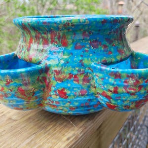 May include: A ceramic strawberry pot with a vibrant turquoise glaze speckled with red and green. The pot has a bulbous top and three side pockets for planting. The pot is sitting on a wooden surface.