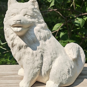 May include: A white ceramic Chow Chow dog figurine, sitting with its tongue out. The sculpture has detailed fur texture and is positioned on a wooden surface, with a blurred green background.