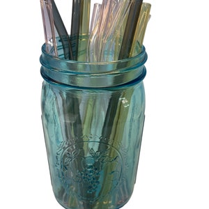 May include: A blue glass jar with a grape design filled with a variety of glass drinking straws in clear, green, and black.