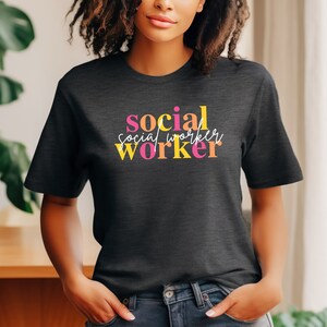 Può includere: Una maglietta grigia scura con la scritta "social worker" stampata in rosa, arancione e giallo.