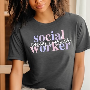 Può includere: Una maglietta grigia scura con la scritta "social worker" stampata in lettere rosa e viola.