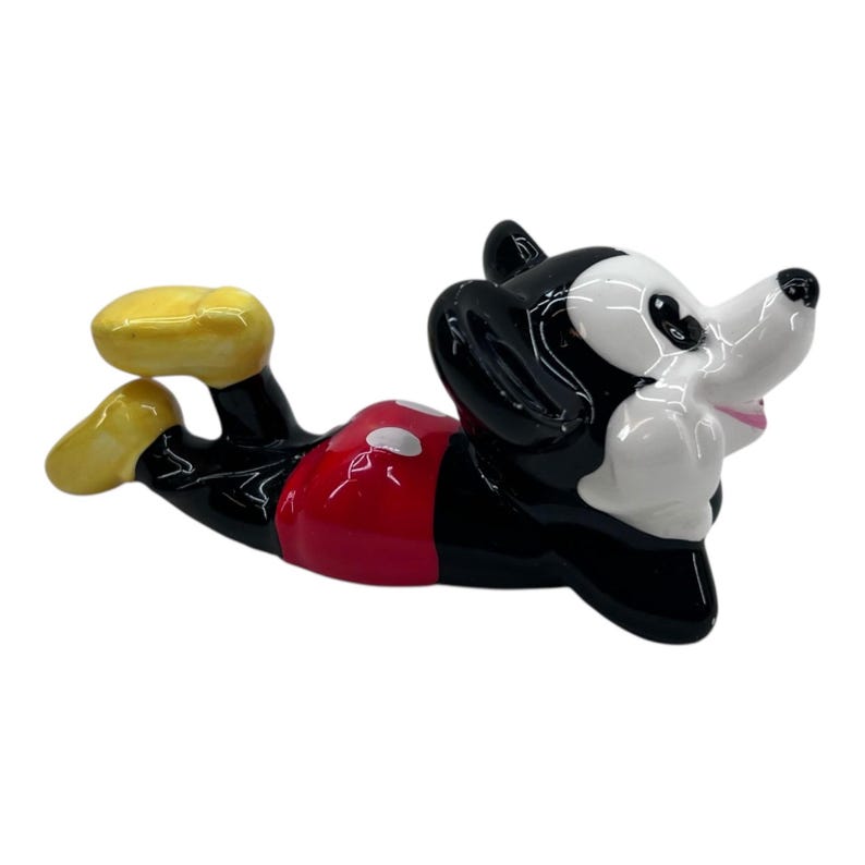 May include: A ceramic figurine of Mickey Mouse in a relaxed pose. The figure is primarily black, with a white face and red shorts with white polka dots. Yellow shoes complete the iconic look.