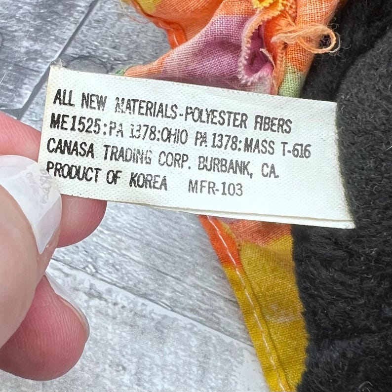 Puede incluir: Primer plano de una etiqueta blanca con texto negro, incluyendo "ALL NEW MATERIALS-POLYESTER FIBERS". La etiqueta est&aacute; junto a una tela colorida con tonos naranja, amarillo y rosa y una tela gris oscuro.