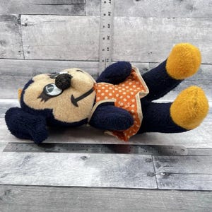 May include: A vintage plush toy in the shape of a cartoon character. The toy has a tan face, navy blue body, and orange feet. It wears an orange and white polka dot dress. The toy is lying on its back.