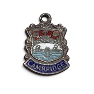 May include: A silver-toned metal charm shaped like a shield with an enamel crest. The design features a red top section, a white middle with three small figures, and a blue base banner with the word CAMBRIDGE in silver capital letters.