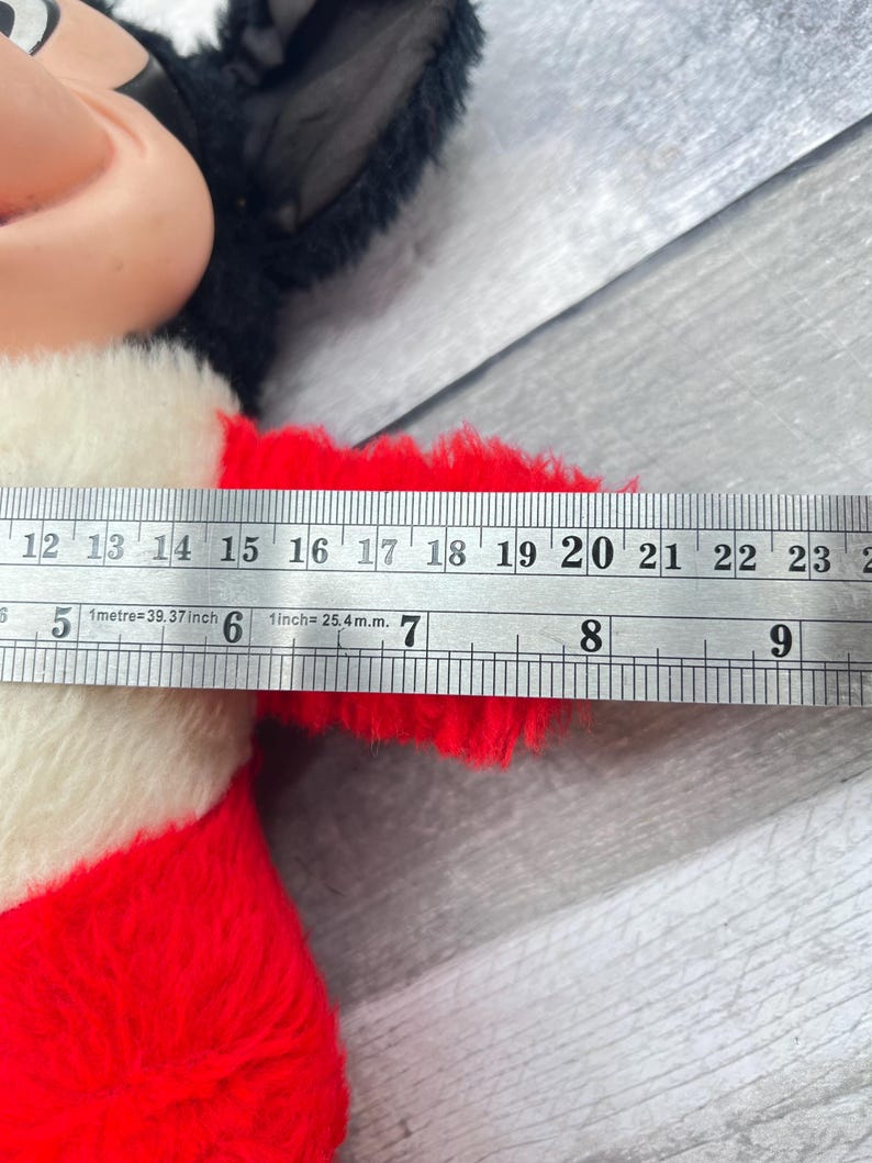 May include: A vintage Mickey Mouse plush toy with black ears, a beige face, and a red and white outfit. The toy's arm measures approximately 18 cm in length, as indicated by a ruler.