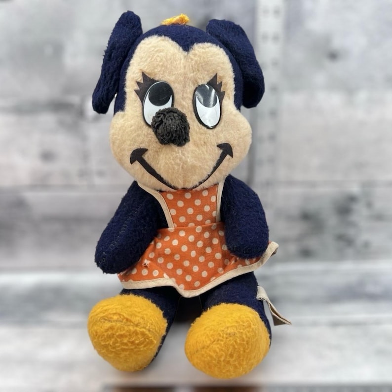 May include: Vintage plush doll featuring a smiling cartoon character with a navy blue head, ears, and arms. The doll has a tan face, black nose, and large eyes. It wears an orange dress with white polka dots and yellow feet.