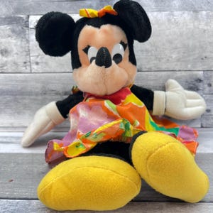 Puede incluir: Mu&ntilde;eca de peluche de Minnie Mouse con orejas negras, un lazo amarillo y un vestido floral colorido. La mu&ntilde;eca tiene guantes blancos y zapatos amarillos grandes. La mu&ntilde;eca est&aacute; sentada sobre una superficie de madera gris.