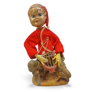 May include: A vintage figurine of a seated figure in a red outfit with gold trim, playing a small brown harp. The figure has a wooden face, blue eyes, and a red hat. The base is a mottled brown and tan color.