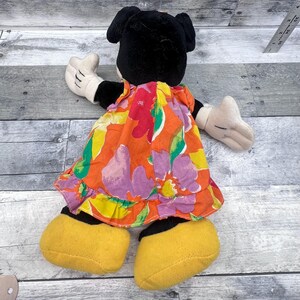 Puede incluir: Mu&ntilde;eca de peluche de Minnie Mouse, vista desde atr&aacute;s, con un vestido estampado floral colorido. La mu&ntilde;eca tiene orejas negras, guantes blancos y zapatos amarillos. El vestido presenta estampados florales naranjas, rosas, amarillos y verdes.