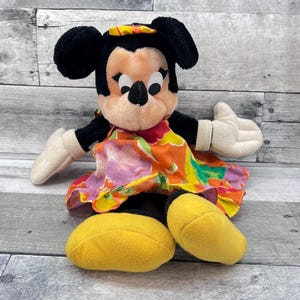Puede incluir: Mu&ntilde;eca de peluche de Minnie Mouse con orejas negras, cara beige y guantes blancos. Lleva un vestido floral colorido con detalles en naranja, rosa y amarillo, y zapatos amarillos. La mu&ntilde;eca est&aacute; sentada.