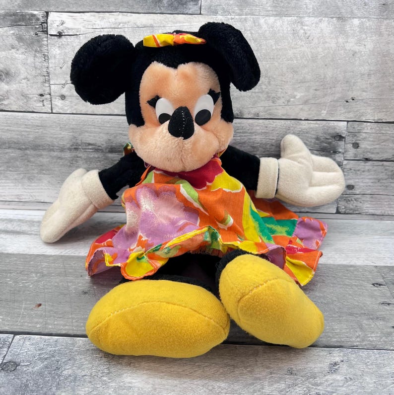 Puede incluir: Mu&ntilde;eca de peluche de Minnie Mouse con orejas negras, cara beige y guantes blancos. Lleva un vestido floral colorido con detalles en naranja, rosa y amarillo, y zapatos amarillos. La mu&ntilde;eca est&aacute; sentada.