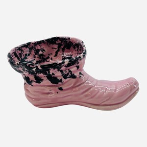 May include: A pink ceramic boot-shaped planter with a black and pink speckled rim. The boot has a curved toe and a textured surface. The planter is designed to hold small plants or decorative items.