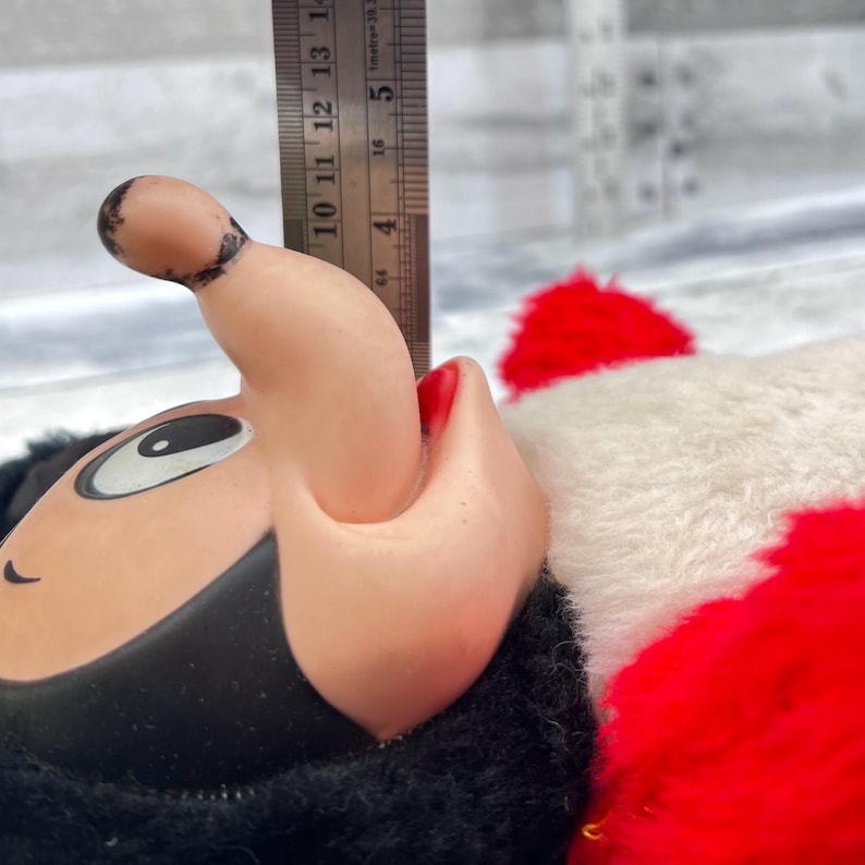 May include: Close-up of a vintage plush toy head with a black hat and face, a pink nose and mouth, and a white eye. The toy has a red and white fluffy body. A ruler is placed next to the toy.