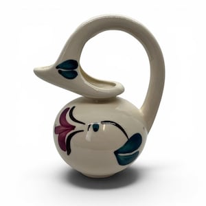 May include: A cream-colored ceramic pitcher shaped like a swan with a long, curved handle forming the neck. The body features hand-painted floral motifs in dark teal and purple, with matching teal accents on the swan's head and beak area.