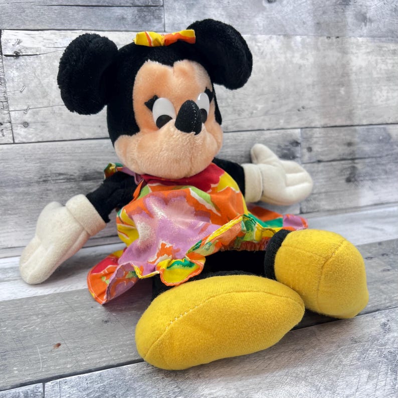 Puede incluir: Mu&ntilde;eca de peluche de Minnie Mouse con orejas negras, un lazo amarillo y un vestido floral colorido. La mu&ntilde;eca tiene guantes blancos y zapatos amarillos. La mu&ntilde;eca est&aacute; sentada sobre una superficie de madera.