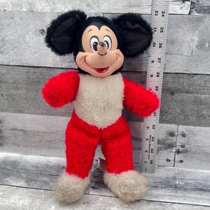 May include: A vintage Mickey Mouse plush toy. The plush has a black head with large ears, a red body, and white accents. A ruler is next to the toy, showing measurements in inches, which are approximately equivalent to cm.