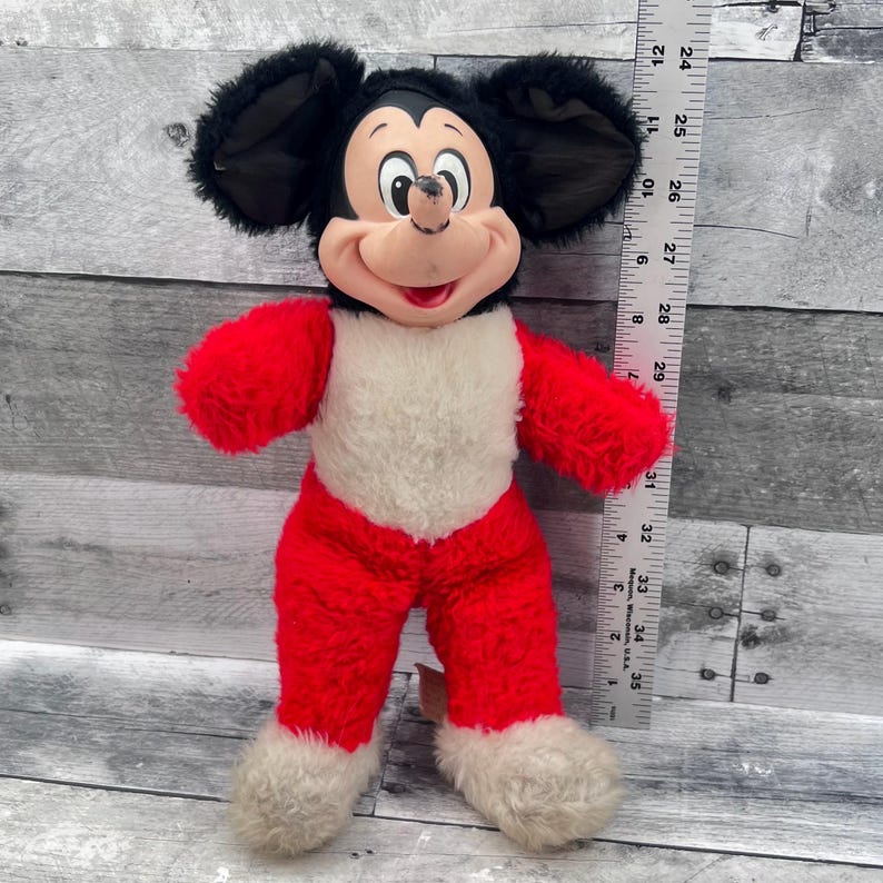 May include: A vintage Mickey Mouse plush toy. The plush has a black head with large ears, a red body, and white accents. A ruler is next to the toy, showing measurements in inches, which are approximately equivalent to cm.