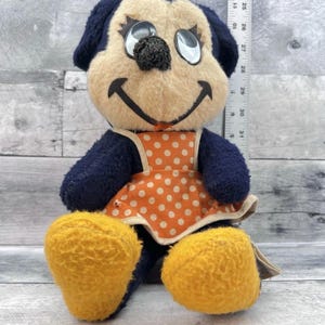 May include: Vintage plush dog toy with a cream-colored face, black nose, and dark blue ears and limbs. It wears an orange polka-dot apron and has yellow feet. The toy is approximately 10 inches tall.